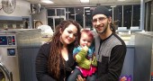 Artist Anna Burke and musician Ryan Stinson, with their daughter, Presley, 18 months, on a recent evening at the laundromat on Dayton Street. Burke and Stinson each moved to Yellow Springs in 2012 from nearby communities. They met here and have begun to build a life in the village, loving the community and navigating the challenges of housing, employment, parenthood and pursuing their art. (Photo by Dylan Taylor-Lehman)