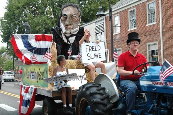 The local effort to erect an “over-life-size” bronze statue of Wheeling Gaunt  made a splash at the Yellow Springs Fourth of July parade last summer. Project steering committee member Dave Neuhardt, president of the the Yellow Springs Historical Society, is behind the tractor wheel. Visible on board the float, which featured a papier-maché depiction of Gaunt’s head, are Malaya Booth and Bob Huston. (Archive photo by Diane Chiddister)