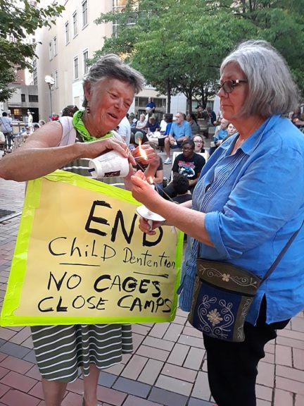 Villager Pat Dewees, left, lights the candle of retired Rev. Lynn Sinnott, of Cedarville, in Dayton at a vigil held in Dayton's Courthouse Square seeking the closure of immigrant detention camps in the U.S. (Photo by Carol Simmons)